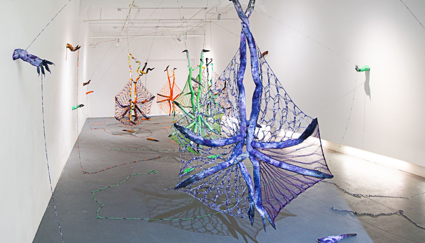 Installation of soft sculpture shaped in large bat-like wings where the membranes are knitted with beads, hands with nails on the wall pulling rope, and motorized tongues on the floor.