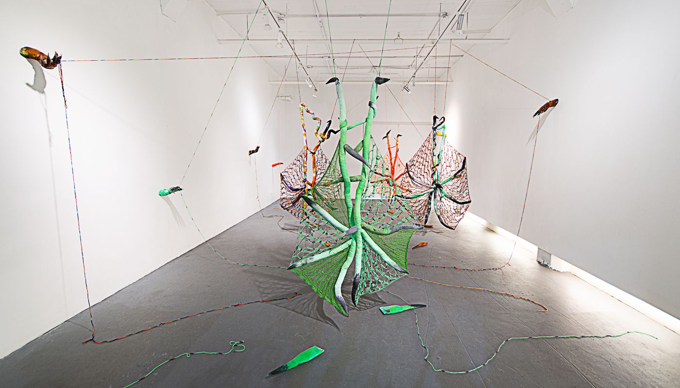 Installation of soft sculpture shaped in large bat-like wings where the membranes are knitted with beads, hands with nails on the wall pulling rope, and motorized tongues on the floor.