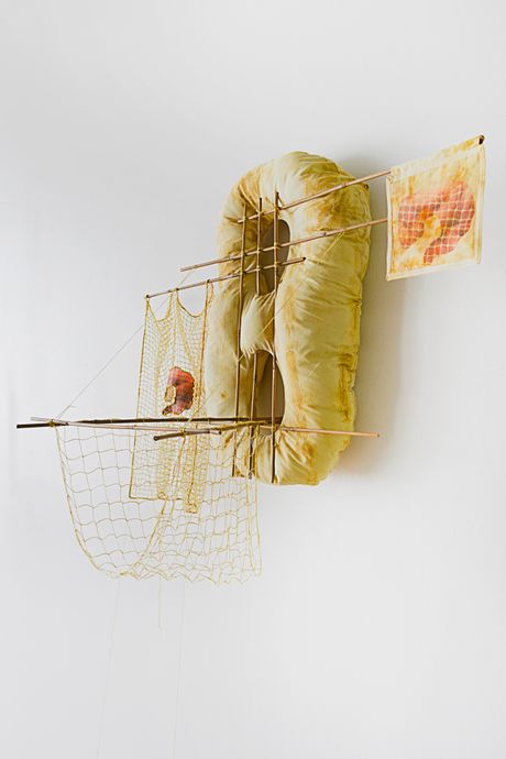 A wall soft sculpture tie dyed in hues of yellow with filet lace and netting elements framed in bamboo that are tied using square lashing knots. Within the filet lace is an embedded gay pornographic image using a sublimation dye technique. Two soft sculptural bananas are attached to the wall sculpture with strings and lies on the floor.
