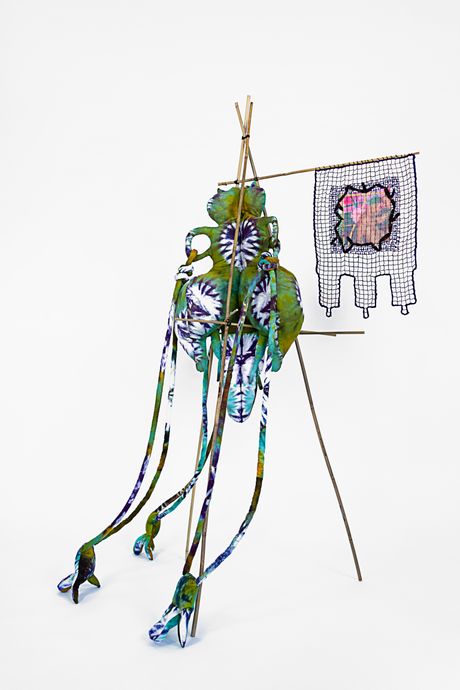 A free standing soft sculpture tie dyed in hues of forest green, primrose yellow, and deep purple with filet lace and netting elements framed in bamboo that are tied using square lashing knots. Within the filet lace is an embedded gay pornographic image using a sublimation dye technique. The soft sculpture element has a main body suspended in the middle of the bamboo frame with holes where a rope-like pieces are tied in a snake knot with talons attached at the end..