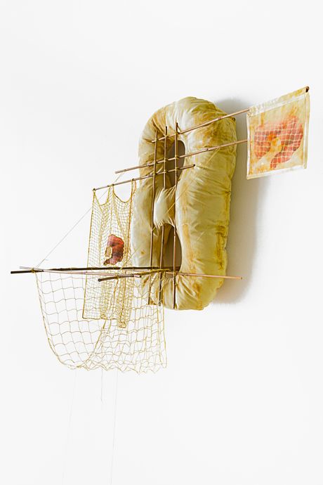 A wall soft sculpture tie dyed in hues of yellow with filet lace and netting elements framed in bamboo that are tied using square lashing knots. Within the filet lace is an embedded gay pornographic image using a sublimation dye technique. Two soft sculptural bananas are attached to the wall sculpture with strings and lies on the floor.