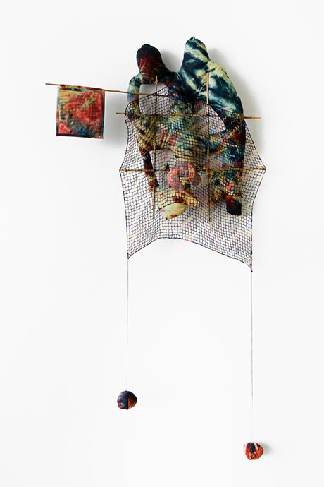 A wall soft sculpture tie dyed in hues of yellow, dark green, and light red with filet lace and netting elements framed in bamboo that are tied using square lashing knots. Within the filet lace is an embedded gay pornographic image using a sublimation dye technique. Two soft sculptural balls are hanging from to the wall sculpture.