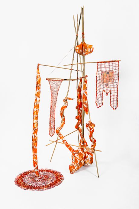 A free standing soft sculpture tie dyed in hues of orange with filet lace and netting elements framed in bamboo that are tied using square lashing knots. Within the filet lace banner is an embedded gay pornographic image using a sublimation dye technique. There is also a cylindrical netting and soft sculptural elements shaped like a luffa gourd.