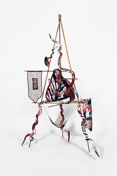 A free standing soft sculpture tie dyed in hues of dark red and dark navy blue with filet lace and netting elements framed in bamboo that are tied using square lashing knots. Within the filet lace is an embedded gay pornographic image using a sublimation dye technique. The soft sculpture element has a main body suspended in the middle of the bamboo frame with tentacles wrapping around the structure.