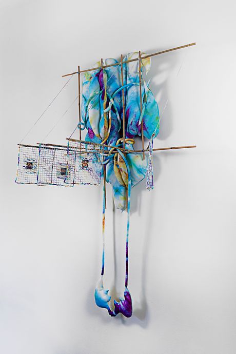 A wall soft sculpture tie dyed in hues of violet, blue, and yellow with filet lace and netting elements framed in bamboo that are tied using square lashing knots. Within the filet lace is an embedded gay pornographic image using a sublimation dye technique.