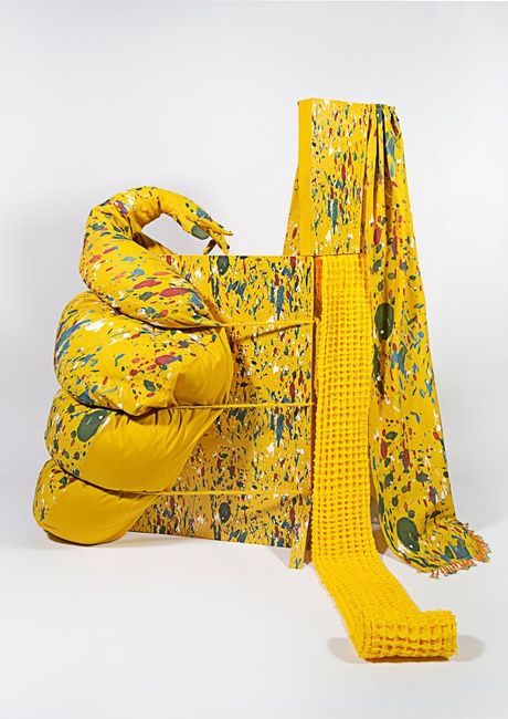 A freestanding sculpture in yellow with spatter patterning using a mixture of fiber techniques (soft sculpture, rope, waffle weave, embroidery, beadwork, and quilting) and wood dyed elements. The shape is bulbous with a hand jutting out and tied with wool rope to a wooden structure. 