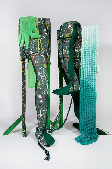 A freestanding sculpture in dark green with spatter patterning using a mixture of fiber techniques (soft sculpture, rope, waffle weave, embroidery, and, beadwork) and wood dyed elements. The sculpture consists of two skinny wooden structures with legs jotting out and a large diaphanous glove opening a zipper. 