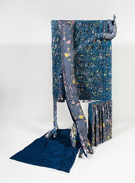 A freestanding sculpture in dark blue with spatter patterning using a mixture of fiber techniques (soft sculpture, trapunto, beadwork, and open knit) and wood dyed elements. The sculpture consists of two rectangles with the smallest supporting the largest on the bottom corner, while a leg supports the other. Two soft sculpture hands wrap around the work, while a rectangular trapunto quilt lays on the ground.
